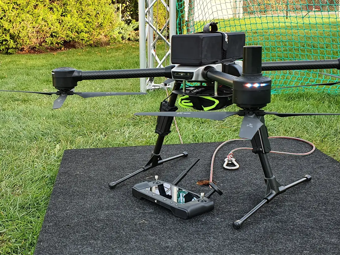 A carbon-fibre quadrotor on a landing pad at a netted flight-test site, with its controller alongside.